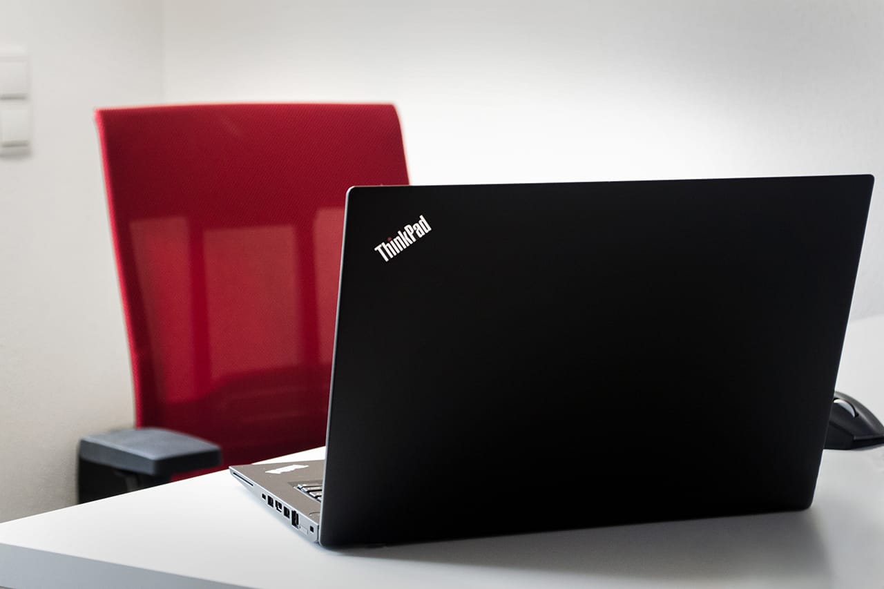 Schwarzer Lenovo ThinkPad Laptop auf weißem Schreibtisch vor rotem Bürostuhl – moderner Arbeitsplatz für IT-Dienstleistungen und Business-Lösungen